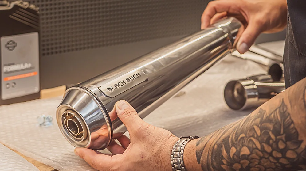 Black Duck Tech Fighter exhaust tail end for Harley-Davidson Softail and Touring on a workbench with technician's hands